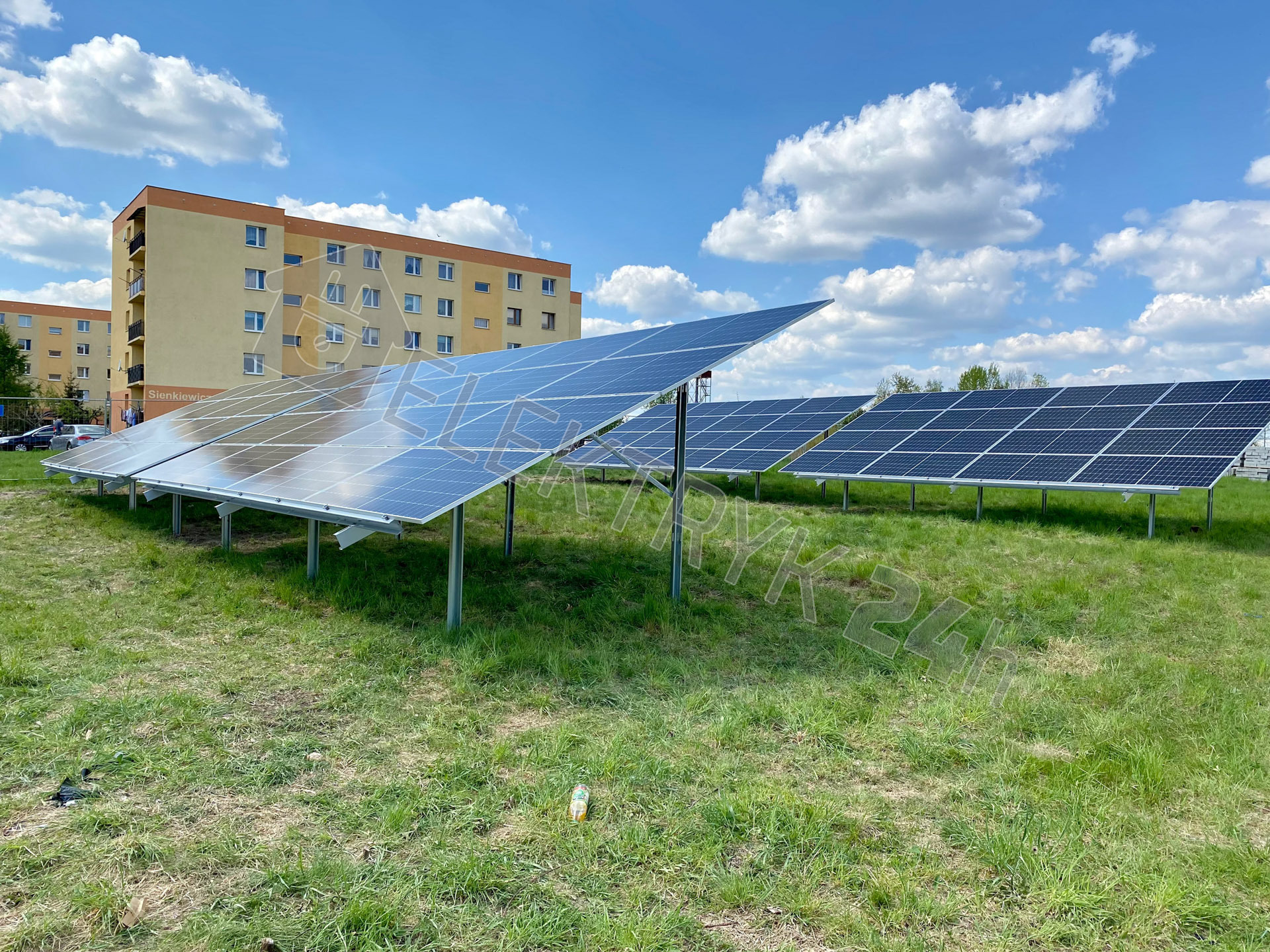 Panele fotowoltaiczne zamontowane na trawiastym terenie, z blokiem mieszkalnym w tle i błękitnym niebem z białymi chmurami.