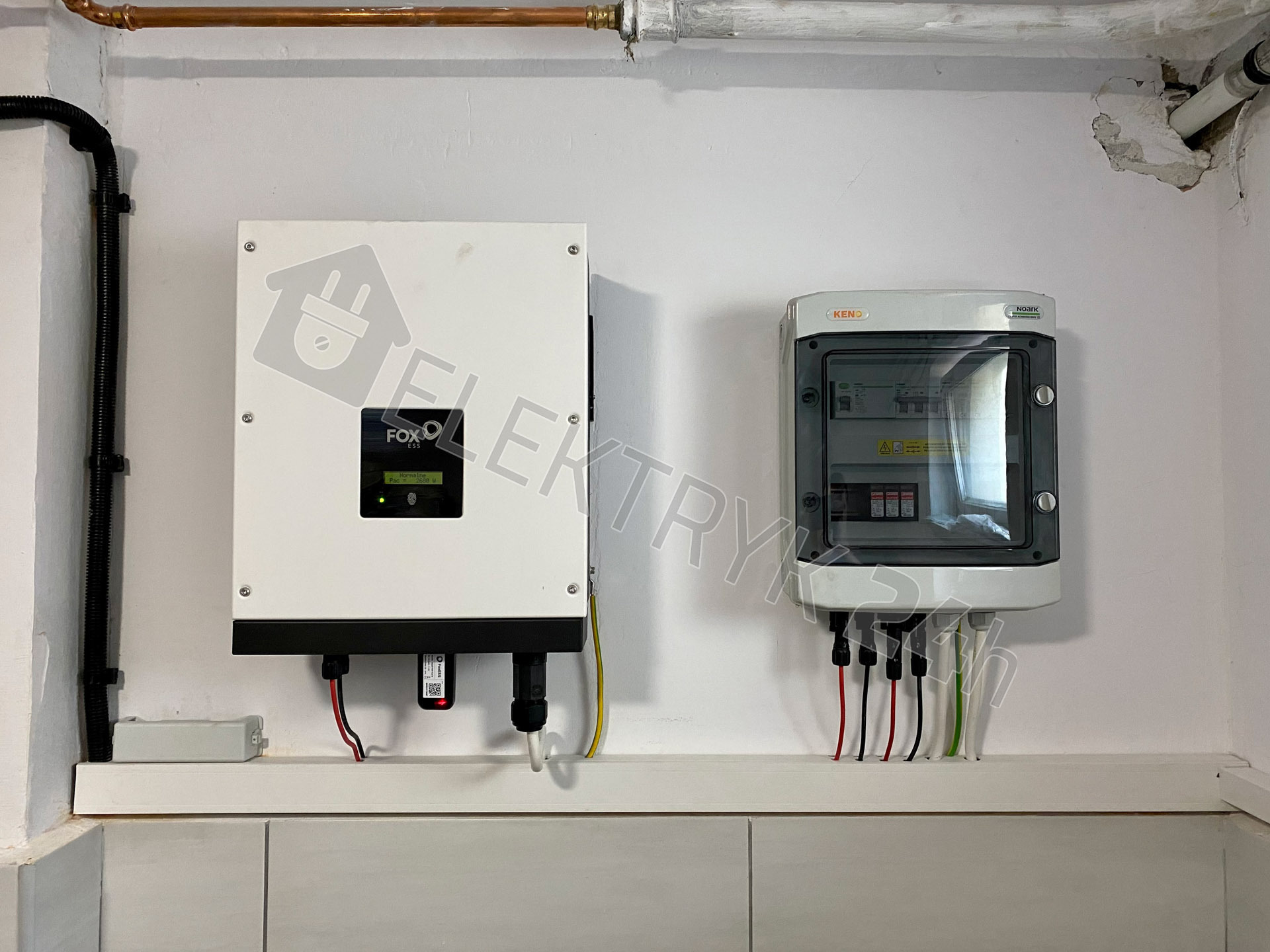 Biała skrzynka inwertera fotowoltaicznego Fox ESS z podłączonymi przewodami i skrzynka rozdzielcza KENO z bezpiecznikami na białej ścianie wewnątrz budynku, widoczne rury instalacji.