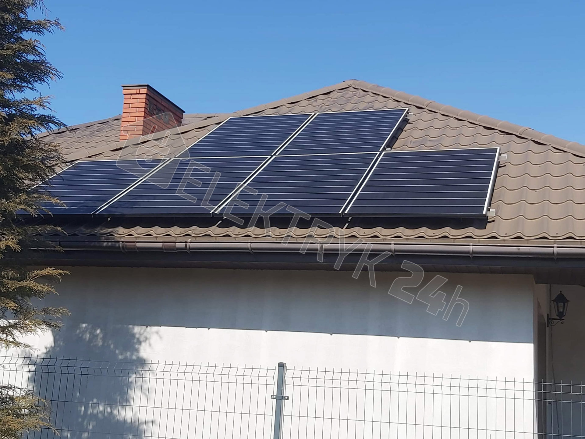 Panele fotowoltaiczne zamontowane na brązowym dachu domu jednorodzinnego z ceglanym kominem, widoczne zza metalowego ogrodzenia.