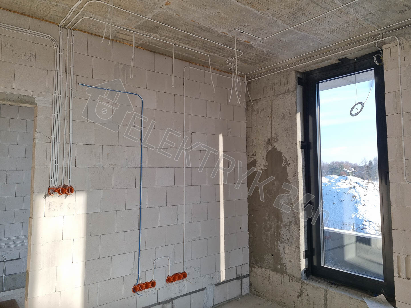 Surowy stan budynku z widocznymi przewodami elektrycznymi w peszlach, prowadzącymi do pomarańczowych puszek instalacyjnych, okno z widokiem na zaśnieżony krajobraz.