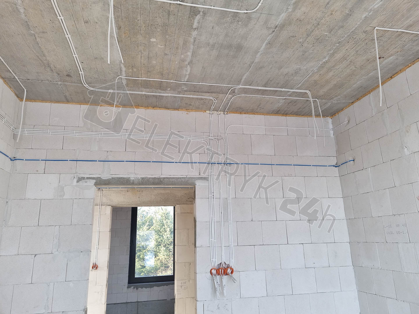 Surowy stan wnętrza z widoczną instalacją elektryczną: białe przewody poprowadzone po suficie i ścianach z betonu komórkowego, wystające kable przy otworze drzwiowym, widok na zewnątrz przez okno.