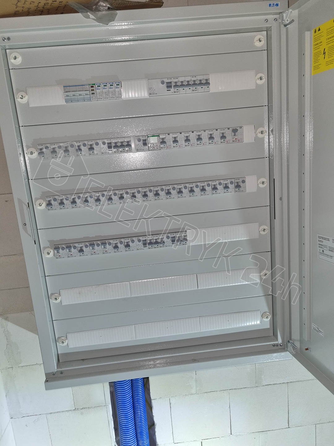 Otwarta szara skrzynka rozdzielcza z zabezpieczeniami elektrycznymi, widoczne niebieskie rury instalacyjne w ścianie z betonu komórkowego.