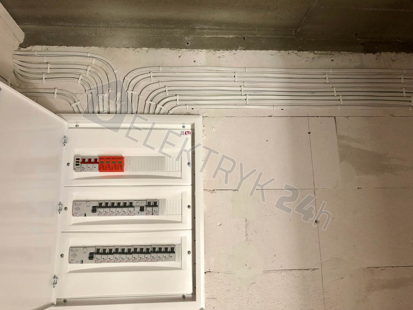 Rozdzielnia elektryczna z otwartymi drzwiczkami, widoczne zabezpieczenia i przewody ułożone w korytkach na ścianie z betonu komórkowego.