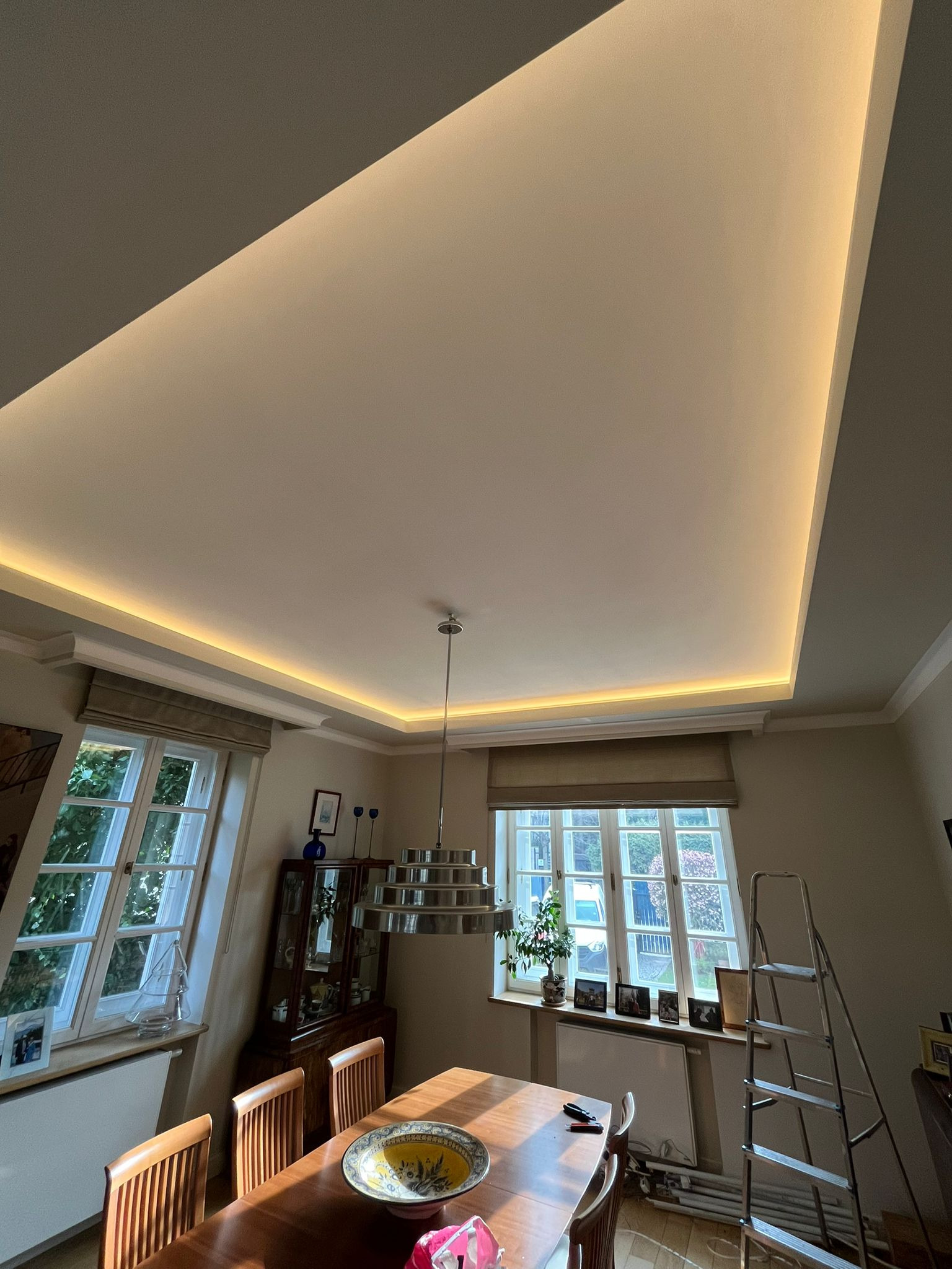 Nowoczesne oświetlenie LED w suficie podwieszanym nad stołem w jadalni, widoczna drabina oparta o ścianę przy oknie.
