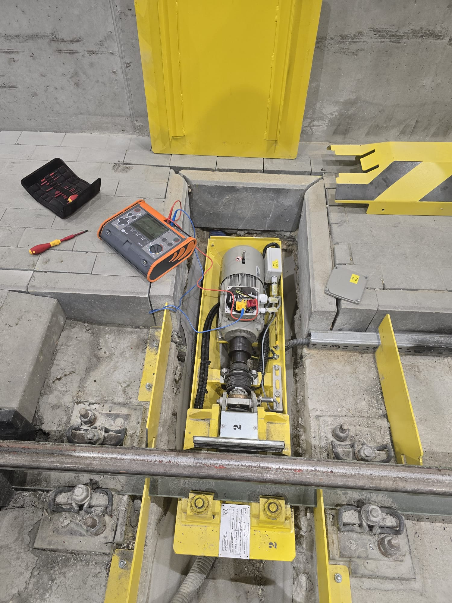 Wykonywanie pomiarów elektrycznych silnika w zagłębieniu posadzki, widoczny miernik, skrzynka z narzędziami i żółte elementy konstrukcyjne, Warszawa.
