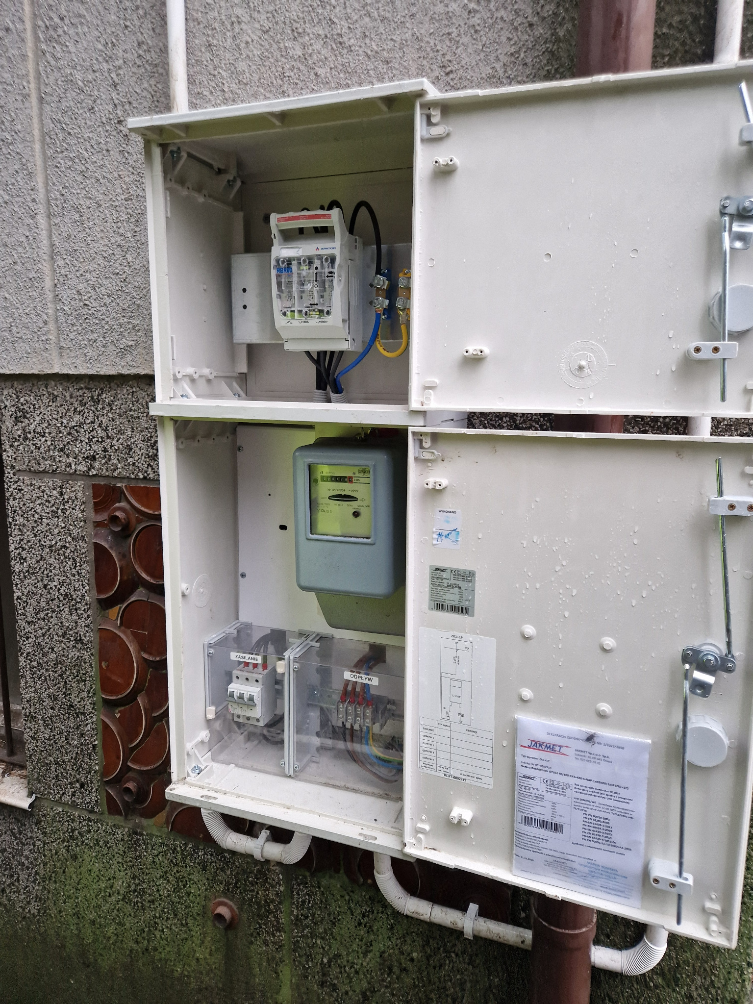 Otwarta skrzynka elektryczna z zamontowanym licznikiem i zabezpieczeniami, widoczne kable, schemat instalacji na drzwiach skrzynki, zewnętrzna ściana budynku z elementami dekoracyjnymi i rurą...