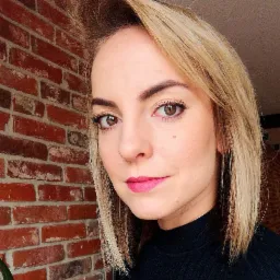 Portret kobiety z blond włosami i różową szminką na tle ceglanej ściany.