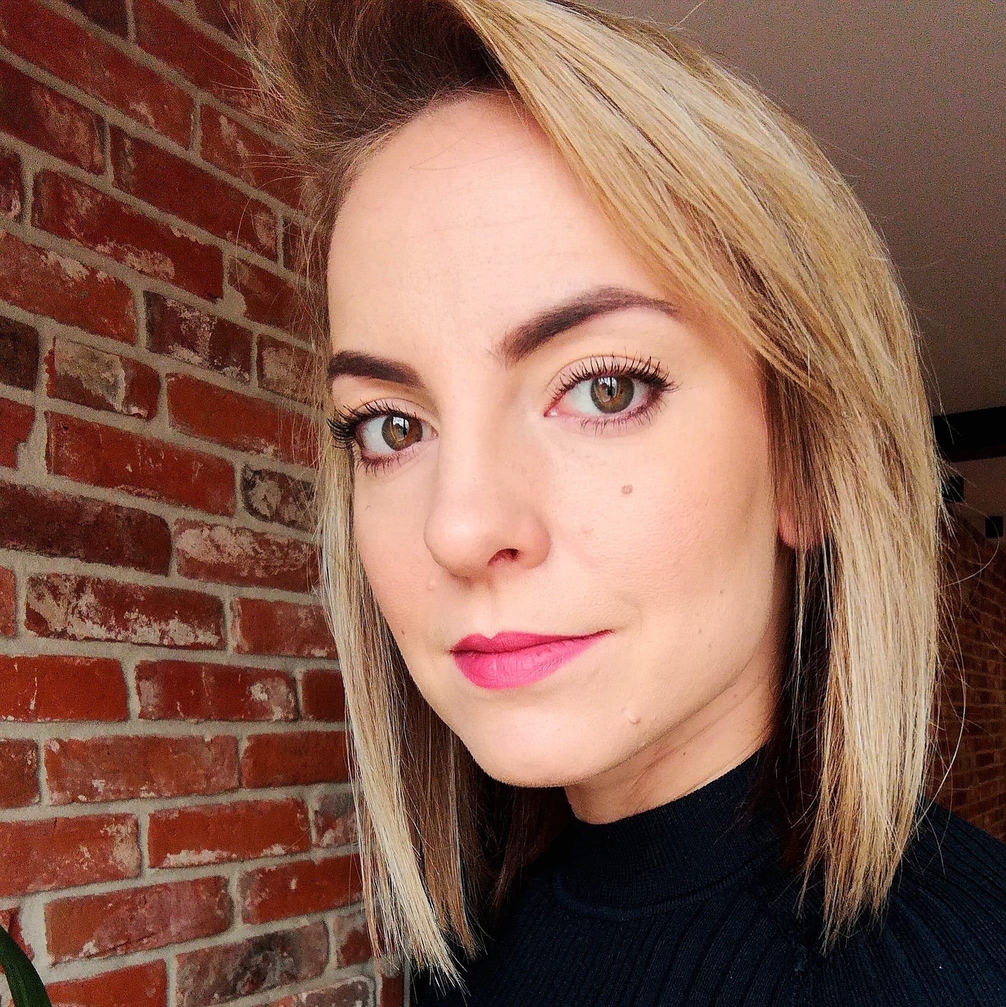 Portret kobiety z blond włosami i różową szminką na tle ceglanej ściany.