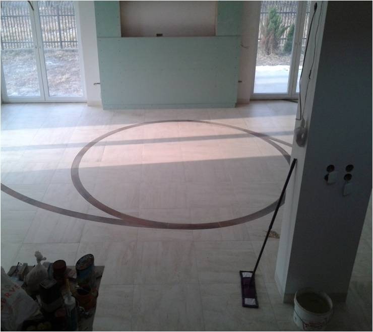 Jasne płytki podłogowe z dekoracyjnym okręgiem z ciemniejszych płytek, widoczne narzędzia i wiadro, ściany w trakcie przygotowania do malowania, duże okna.