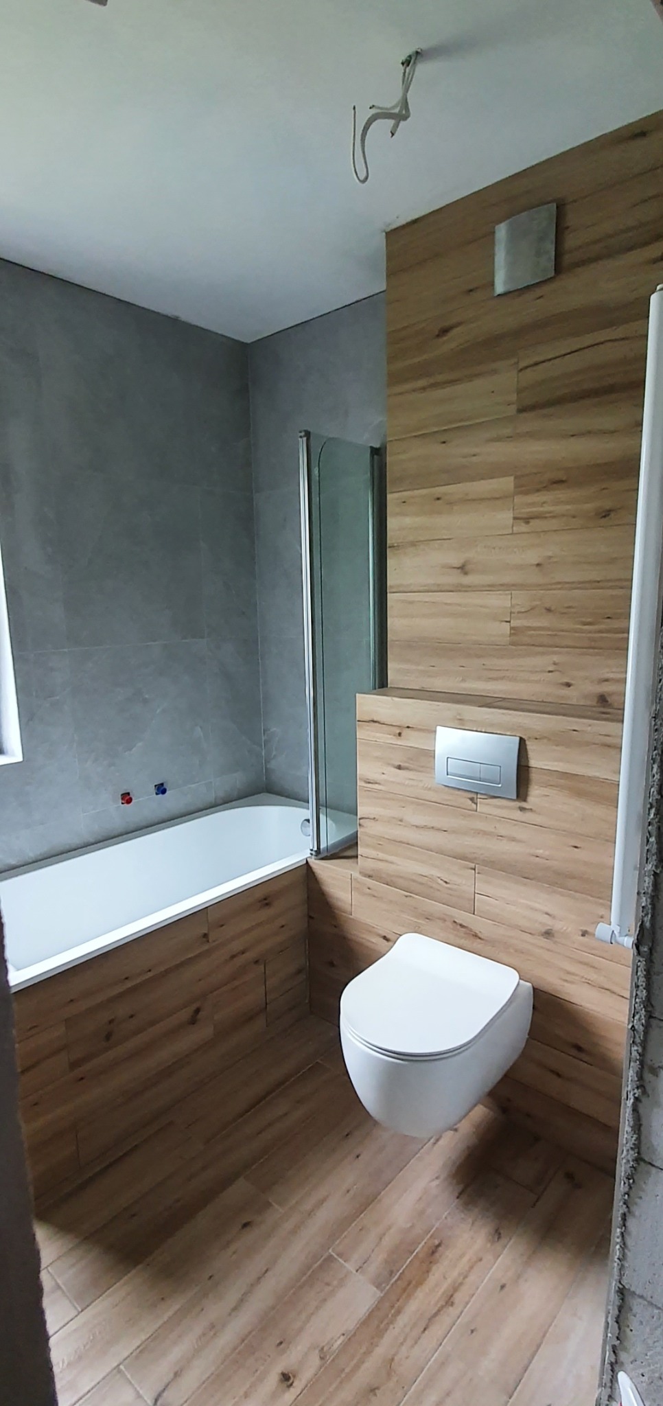 Nowoczesna łazienka z wanną, kabiną prysznicową i podwieszaną toaletą, wykończona płytkami imitującymi drewno i szarym kamieniem, z widoczną instalacją elektryczną na suficie.