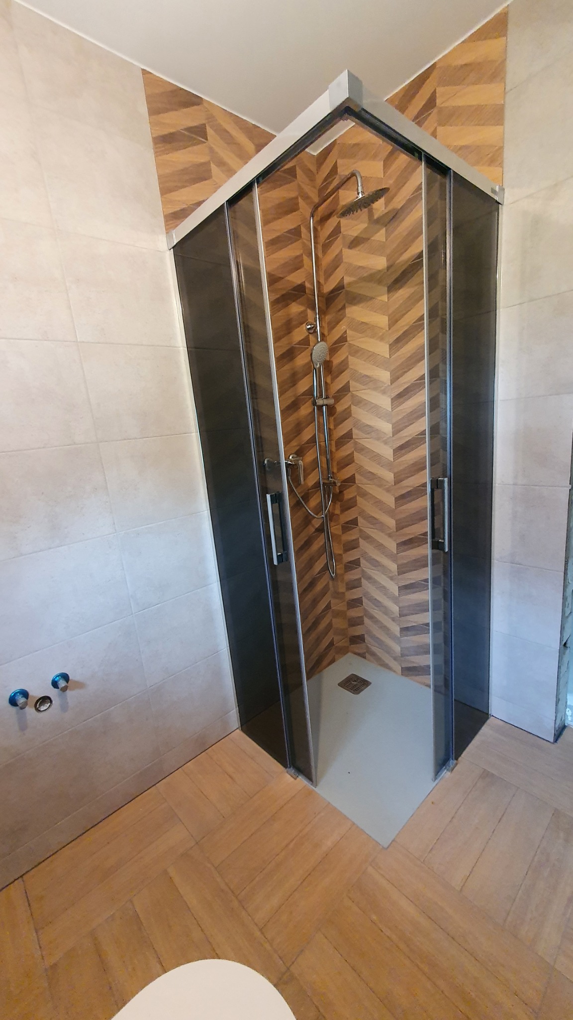 Nowoczesna kabina prysznicowa z ciemnymi ramami i deszczownicą, ściany wyłożone płytkami w jodełkę, widoczne przyłącza wodne na ścianie.