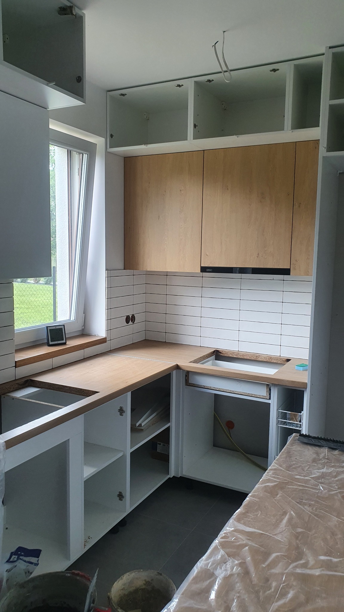 Montaż kuchni: surowe drewniane blaty, białe szafki dolne bez frontów, górne szafki z drewnianymi frontami, białe płytki na ścianie. Widoczne przygotowania do podłączenia instalacji.