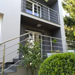 Nowoczesny dom z szarą elewacją, balkonem i schodami zabezpieczonymi balustradami ze stali nierdzewnej; widoczne krzewy i drzewa w otoczeniu.