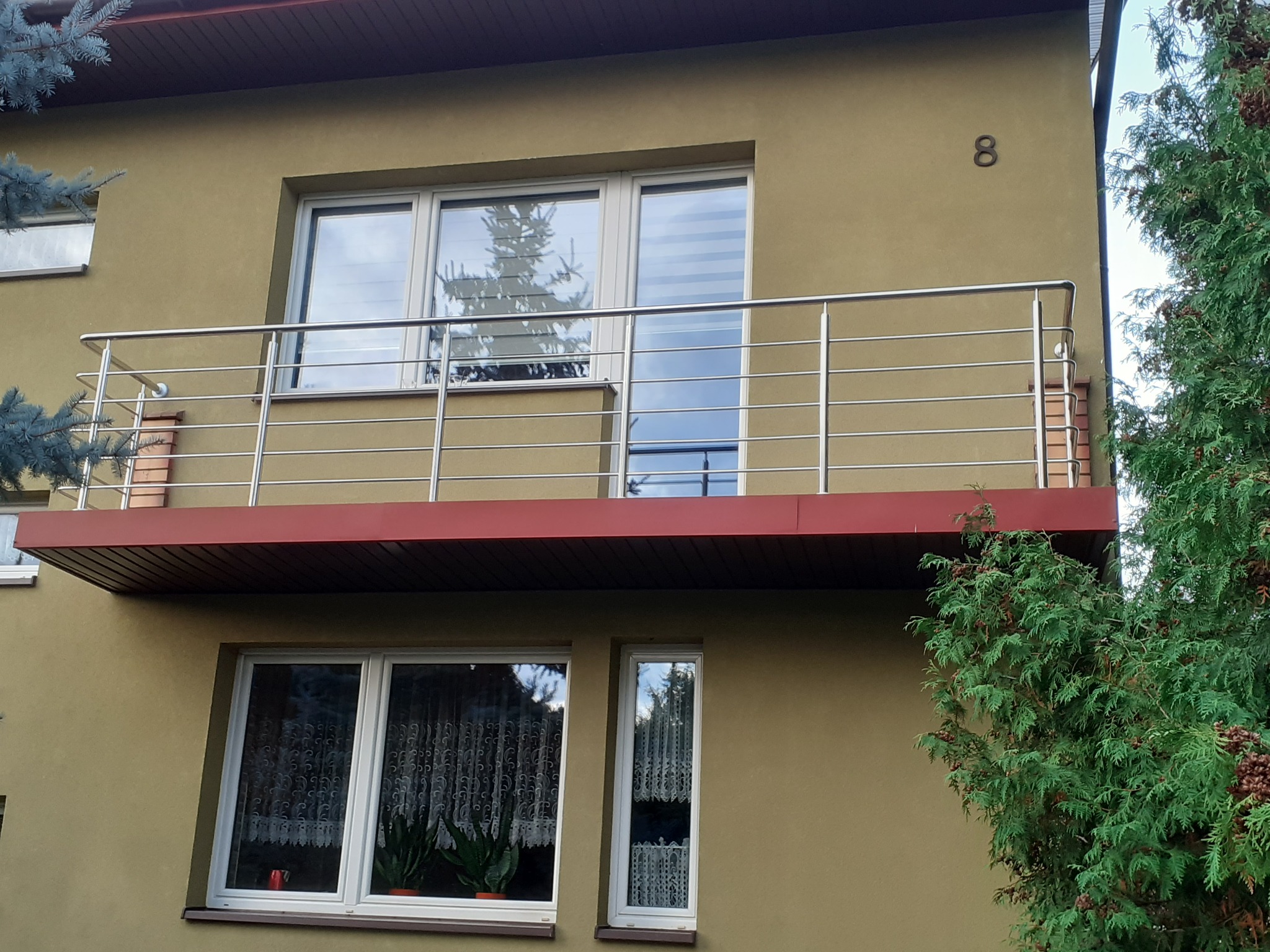 Zewnętrzny widok balkonu z prostą, poziomą balustradą ze stali na tle elewacji budynku w odcieniach beżu, widoczne okna z firankami i roślinami doniczkowymi oraz fragment zielonego drzewa.