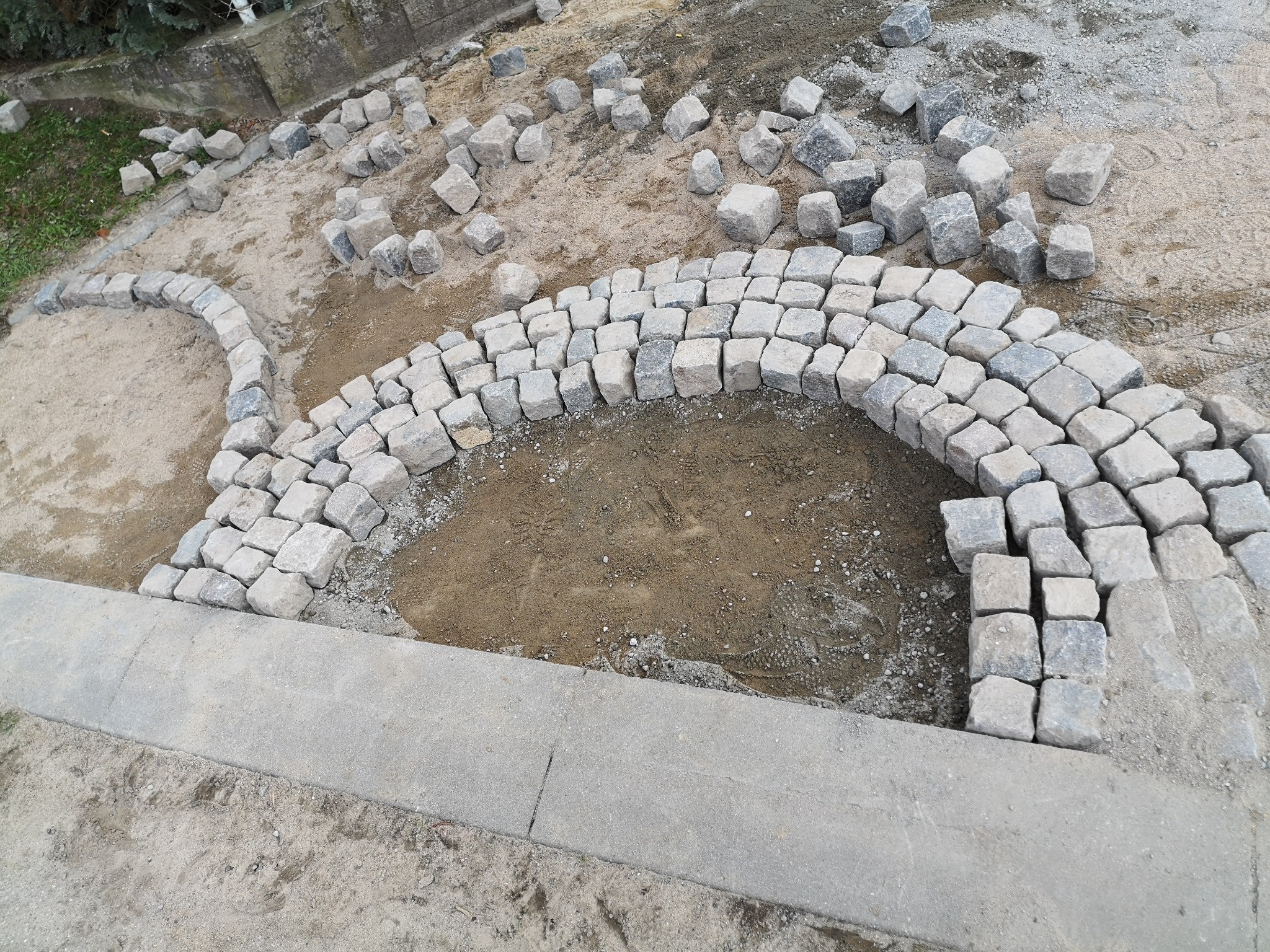 Niedokończony łuk z kostki granitowej na piaskowym podłożu, część kostek ułożona, reszta przygotowana do ułożenia, widoczny betonowy obrzeże.