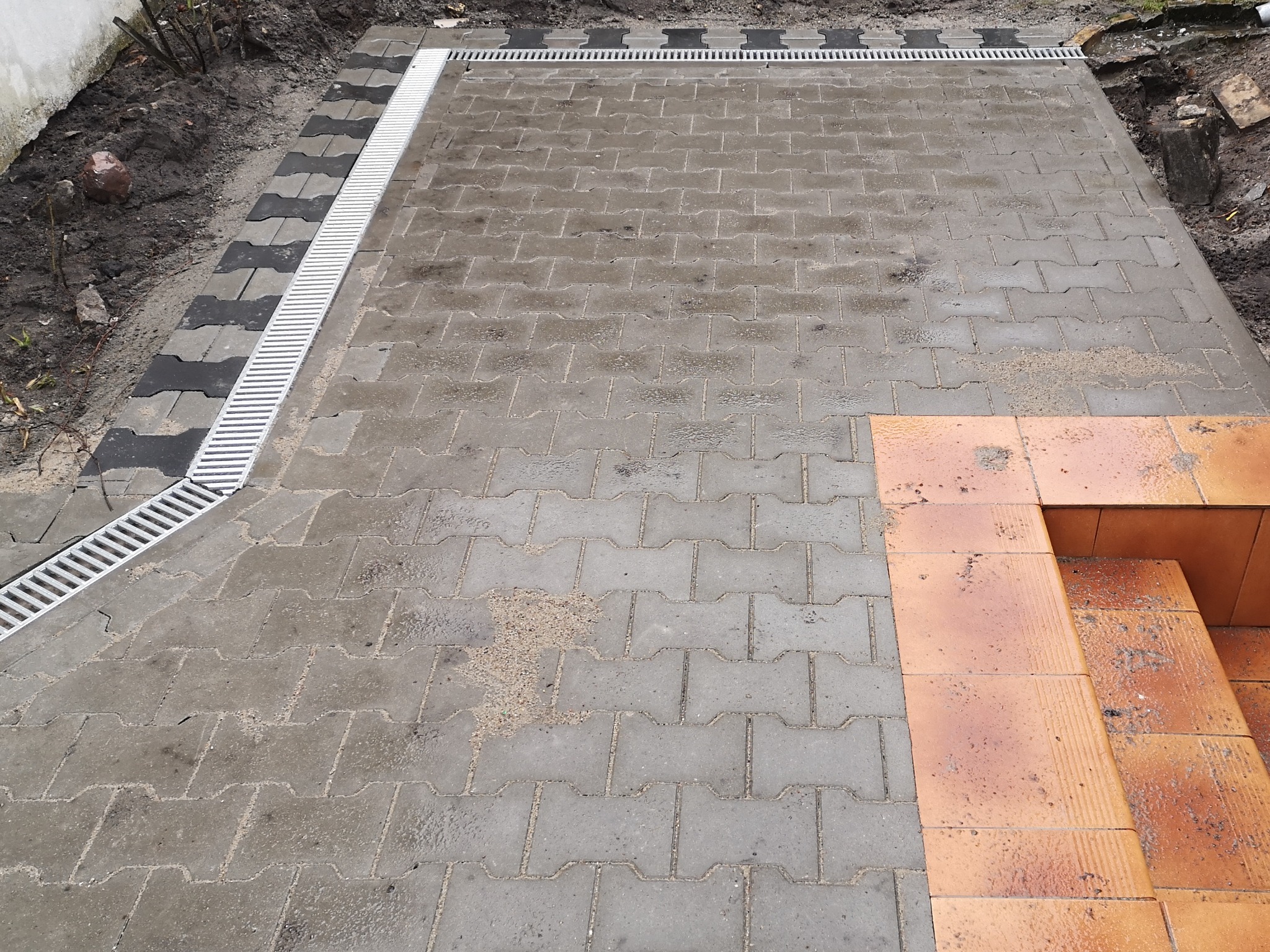 Szara kostka brukowa ułożona w kształt prostokąta z metalowym odpływem liniowym na obrzeżach, sąsiadująca z ceglanymi schodami i ziemią.