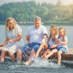 Uśmiechnięta rodzina siedzi na drewnianym pomoście nad jeziorem, chlapiąc wodą nogami w słoneczny dzień. Ojciec z tatuażem na ramieniu robi śmieszną minę.