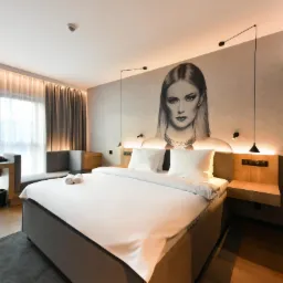 Nowoczesne wnętrze sypialni hotelowej z łóżkiem king-size, biurkiem przy oknie i portretem kobiety na ścianie za łóżkiem.