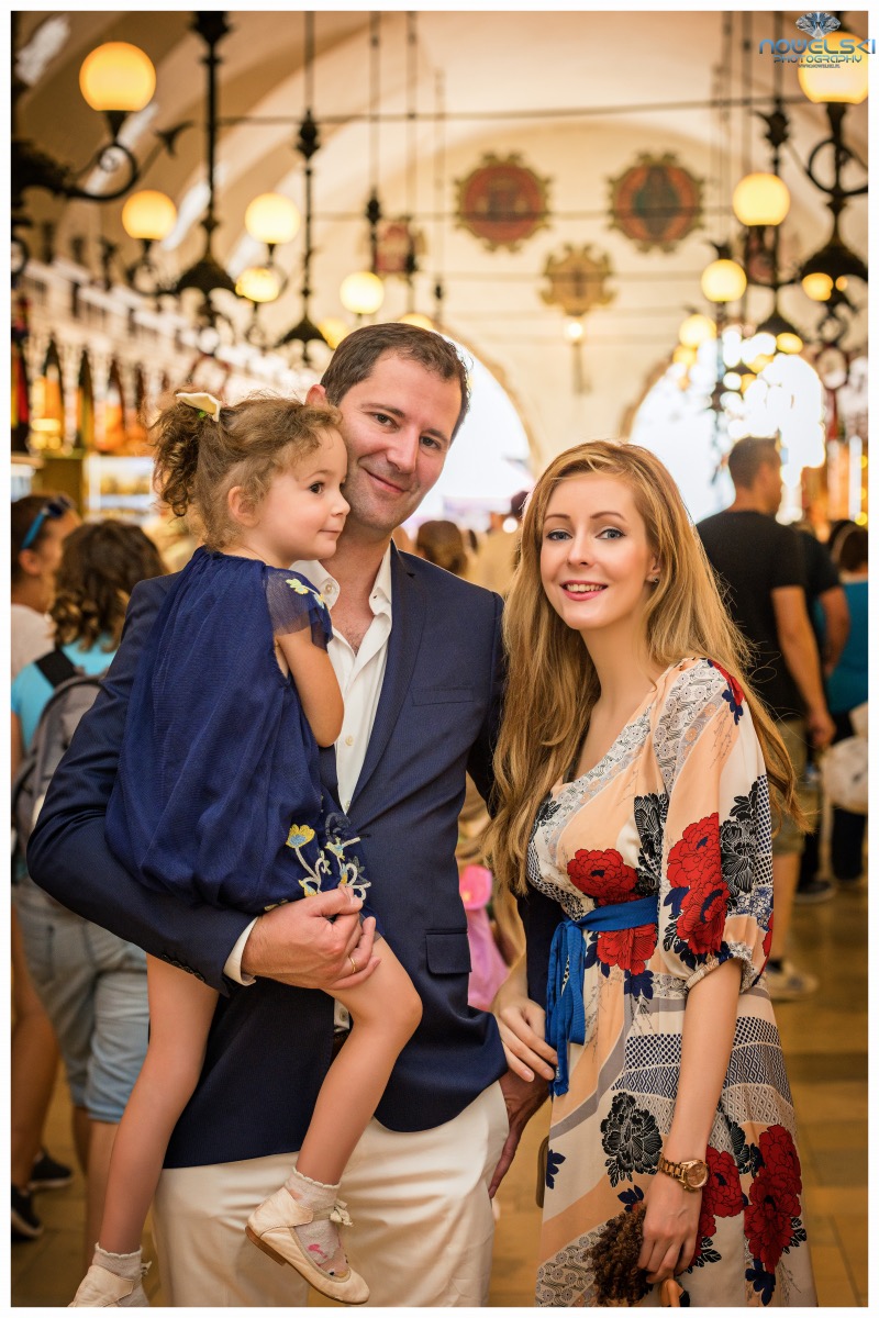 Rodzinne zdjęcie portretowe: ojciec trzyma córkę na rękach, obok stoi matka, w tle zabytkowa hala targowa z ozdobnymi lampami.
