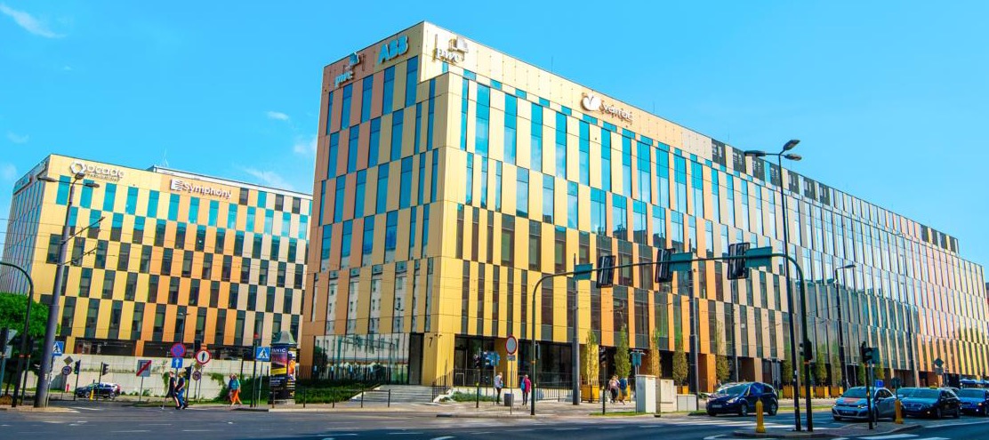 Nowoczesny budynek biurowy z charakterystyczną fasadą w odcieniach żółci i błękitu, z widocznymi logotypami firm ABB, PwC i Symphony na elewacji.