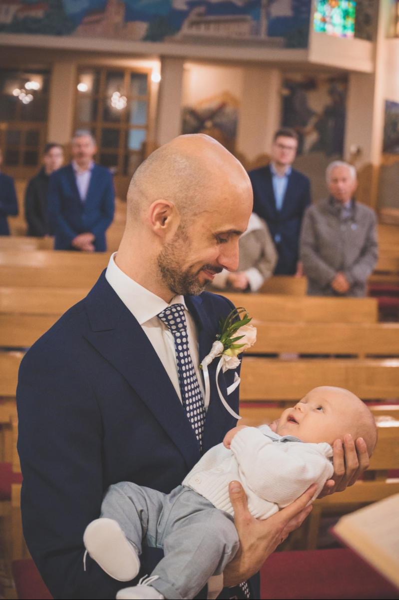 Uśmiechnięty mężczyzna w garniturze z butonierką, trzymający na rękach ubranego niemowlaka w kościele, w tle widoczni goście.