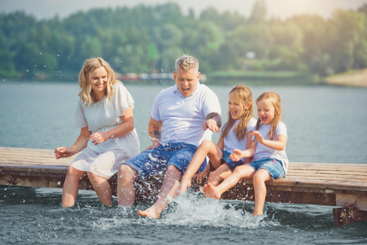 Uśmiechnięta rodzina siedzi na drewnianym pomoście nad jeziorem, chlapiąc wodą nogami w słoneczny dzień. Ojciec z tatuażem na ramieniu robi śmieszną minę.