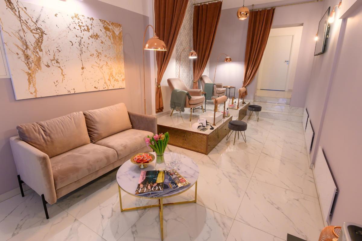 Wnętrze salonu kosmetycznego z beżową sofą, marmurowym stolikiem kawowym z bukietem tulipanów i magazynami Vogue, stanowiskami do manicure z lampami i fotelami w tle, całość utrzymana w pastelowych...