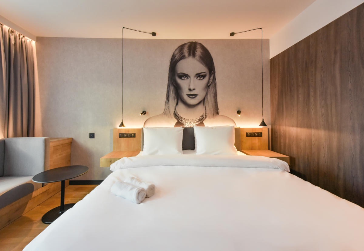 Nowoczesny pokój hotelowy z łóżkiem king-size, białymi ręcznikami i portretem kobiety na ścianie, oświetlony lampkami nocnymi.