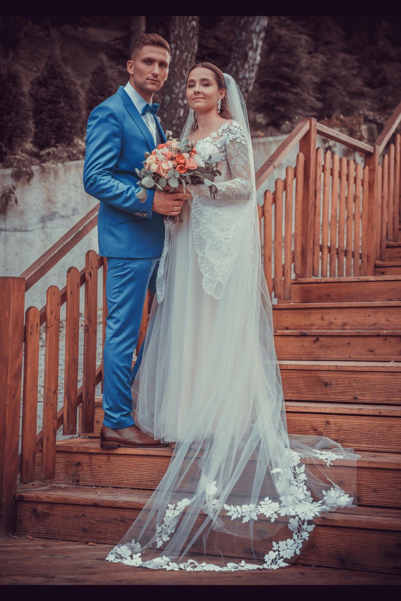 Para młoda na drewnianych schodach: on w niebieskim garniturze z muchą, ona w białej sukni z koronkowymi rękawami i długim welonem, trzyma bukiet z różowych i pomarańczowych kwiatów.