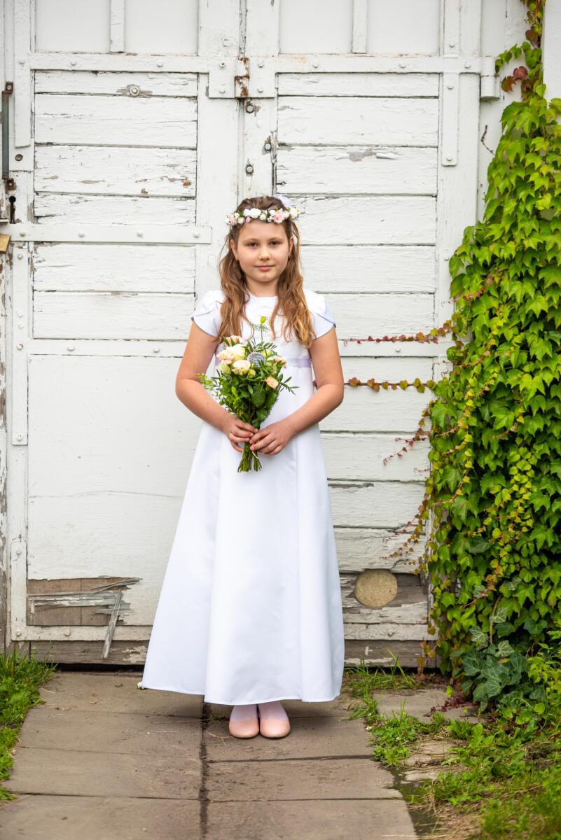 Dziewczynka w białej sukience komunijnej z wiankiem na głowie trzyma bukiet kwiatów na tle starych, białych drzwi obrośniętych bluszczem.
