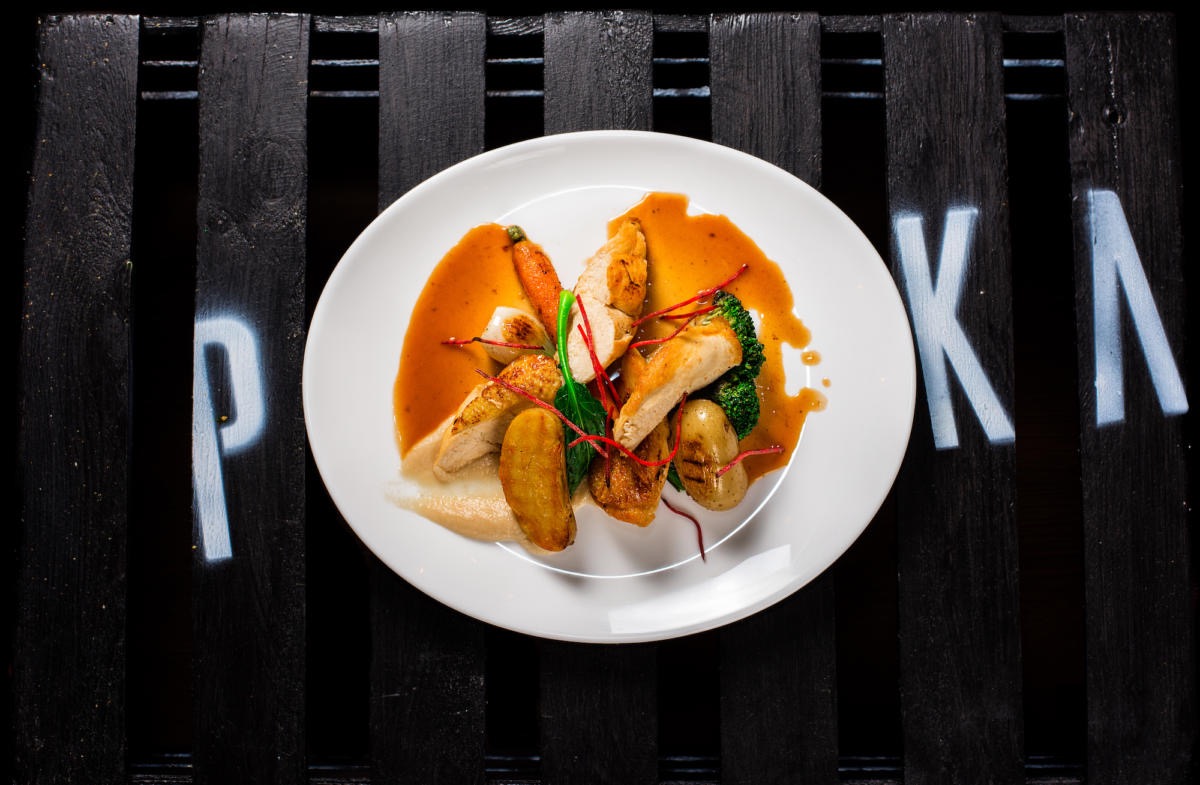 Elegancko podane danie z grillowanym kurczakiem, mini marchewką, brokułem i ziemniakami w sosie na białym talerzu, ułożone na ciemnym drewnianym tle z białymi literami 'P KA'.