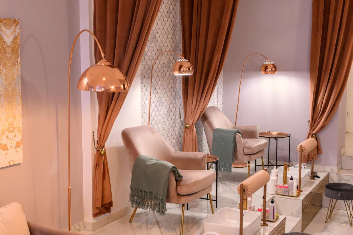Eleganckie wnętrze salonu kosmetycznego z fotelami w odcieniach różu, miedzianymi lampami łukowymi i stanowiskami do manicure z białymi misami.