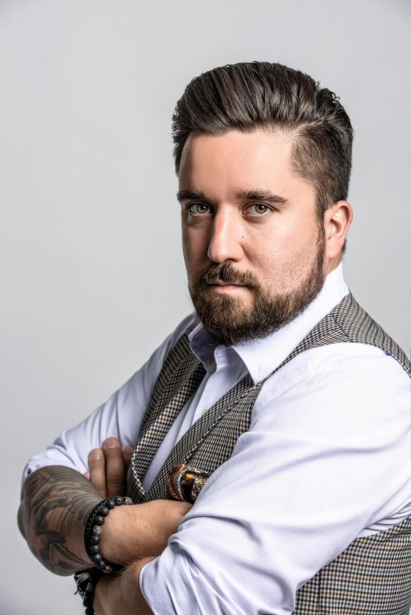 Portret brodatego mężczyzny w koszuli i kraciastej kamizelce, z tatuażem na ramieniu i bransoletkami na nadgarstku, pozującego na jasnym tle.