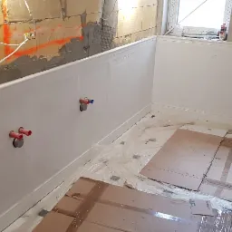 Łazienka w trakcie prac wykończeniowych: białe panele ścienne z dekoracyjnymi wgłębieniami, widoczne rury wystające ze ściany, zabezpieczona folią i kartonami podłoga, fragment surowej ściany...