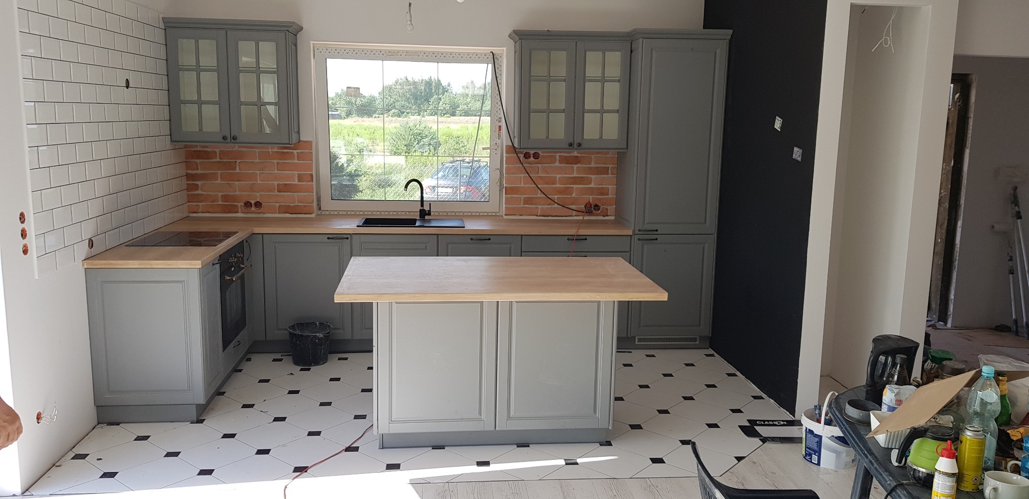 Szara kuchnia z wyspą, ceglanym i białym kafelkowym backsplashiem, drewnianym blatem i czarną armaturą, widoczna przez okno zieleń