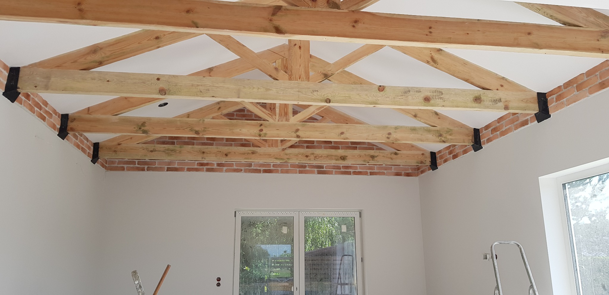 Wyeksponowane drewniane belki stropowe z ceglaną obudową i czarnymi łącznikami w jasnym wnętrzu, widoczne drabiny malarskie.