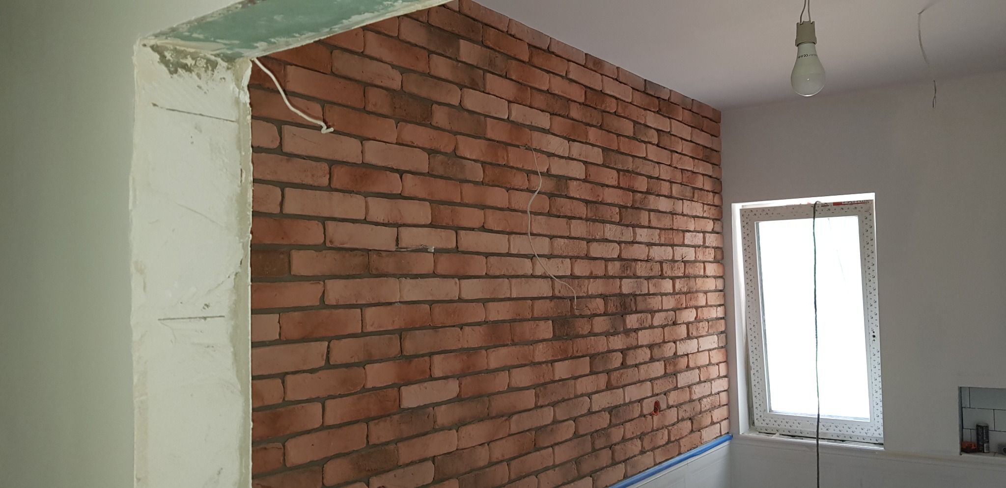 Ściana wyłożona cegłą klinkierową w trakcie prac wykończeniowych, widoczne przewody elektryczne i niezakończone obramowanie drzwi.