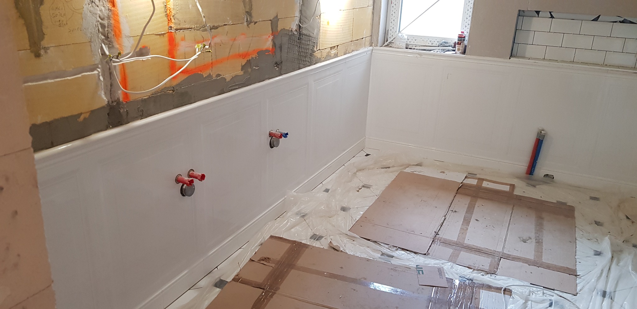 Łazienka w trakcie prac wykończeniowych: białe panele ścienne z dekoracyjnymi wgłębieniami, widoczne rury wystające ze ściany, zabezpieczona folią i kartonami podłoga, fragment surowej ściany...