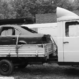 Czarno-białe zdjęcie białego samochodu dostawczego z otwartą platformą, na której przewożony jest wrak czarnego samochodu osobowego, zabezpieczony pasami transportowymi. W tle budynek gospodarczy...