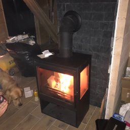 Dr. INSTAL Instalacje Sanitarne - Czarny piec wolnostojący z palącym się w środku drewnem, podłączony do komina z czarnej blachy. W tle ściana wyłożona czarnym kamieniem oraz prowizoryczne wykończenie wnętrza. Na podłodze widoczne...