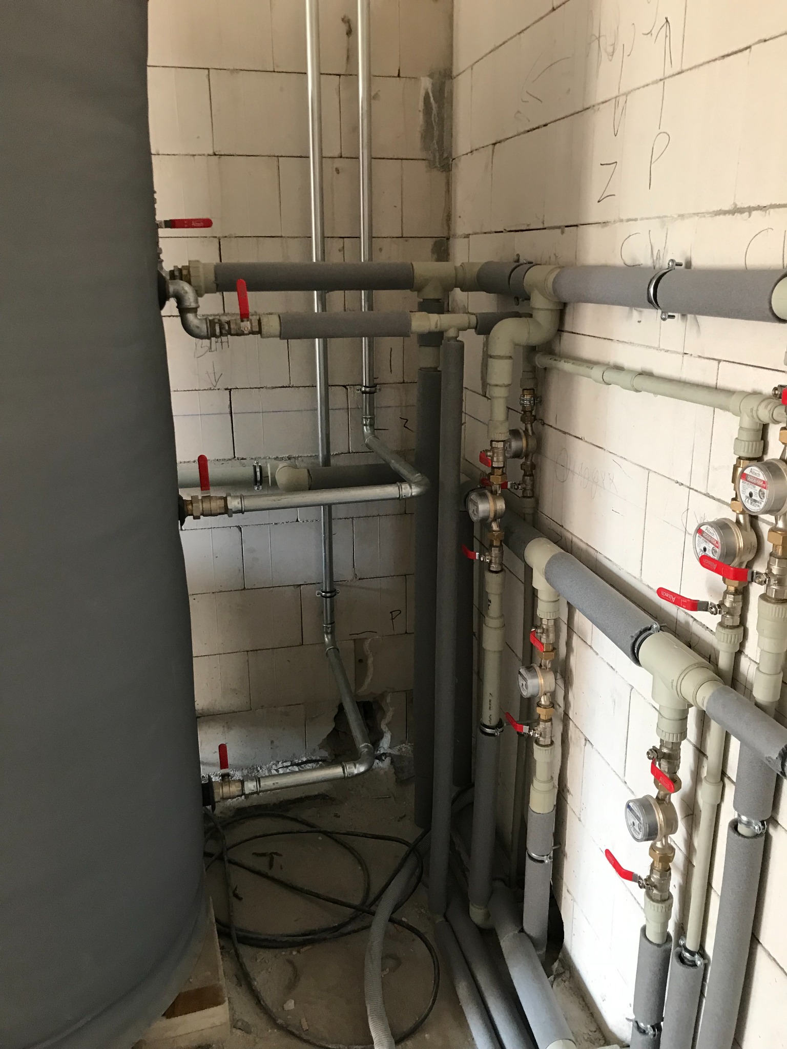 Szare rury instalacji wodnej i grzewczej z czerwonymi zaworami, częściowo zaizolowane, prowadzone wzdłuż ściany z białych bloczków, widoczne liczniki wody.