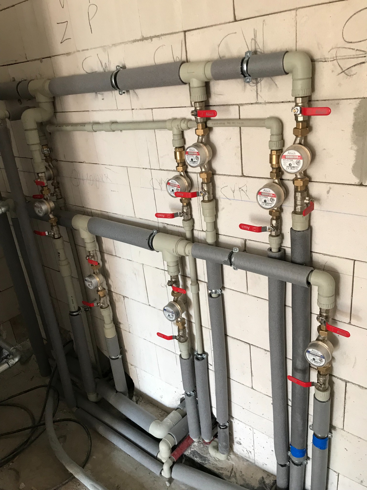 Instalacja wodna z rurami w izolacji, zaworami z czerwonymi uchwytami i licznikami wody zamontowana na ścianie z białych cegieł. Widoczne napisy na ścianie. Część rur zabezpieczona taśmą.