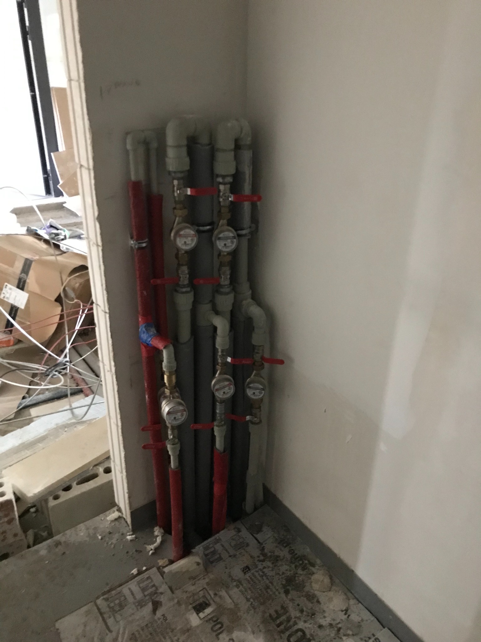 Pionowe rzędy szarych i czerwonych rur instalacji hydraulicznej z zaworami i licznikami, zamontowane przy ścianie w surowym stanie deweloperskim.