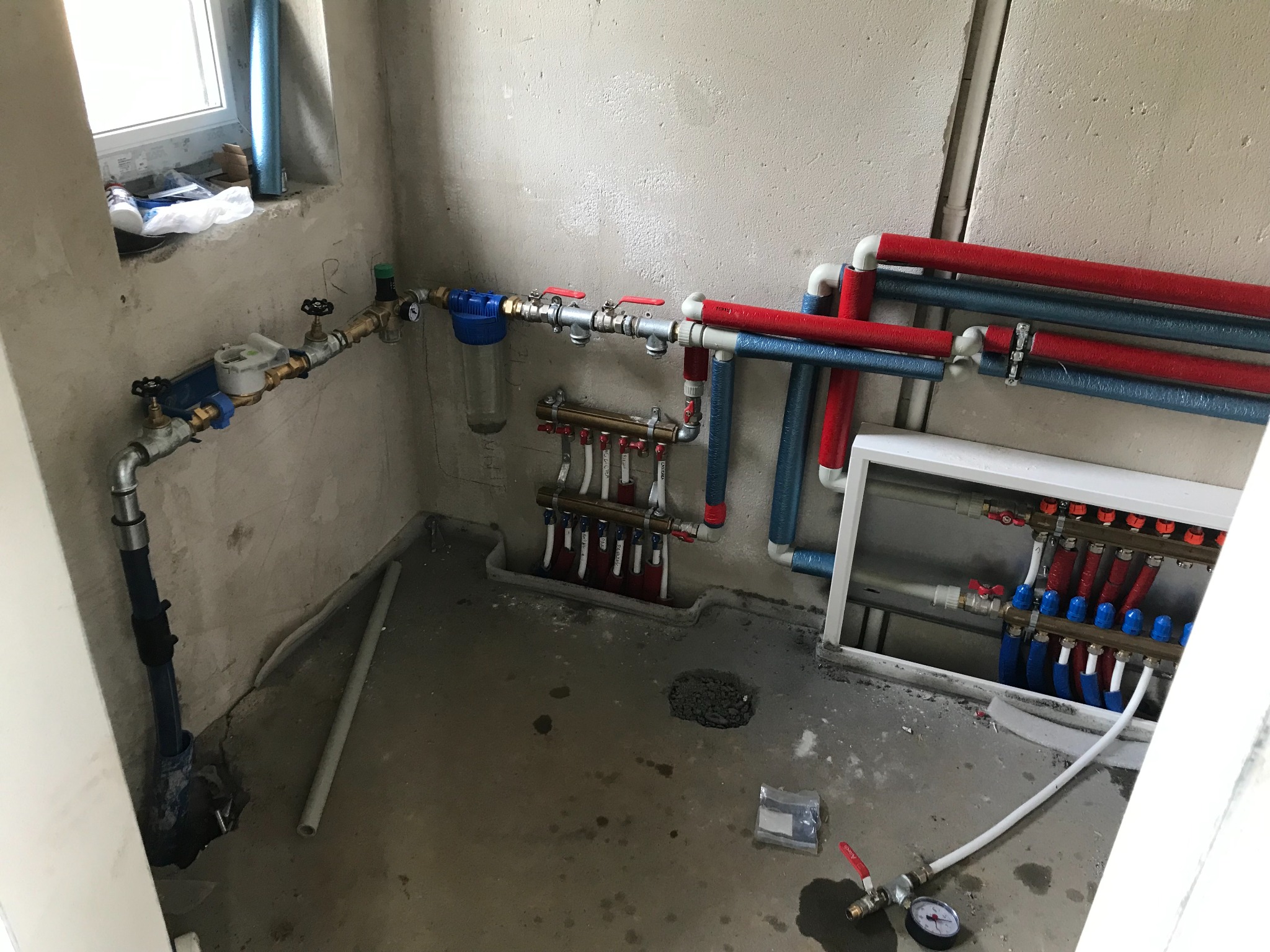 Rozdzielacz ogrzewania podłogowego z rurami w izolacji czerwonej i niebieskiej, wodomierz z filtrem mechanicznym, manometr na podłodze w surowym pomieszczeniu.