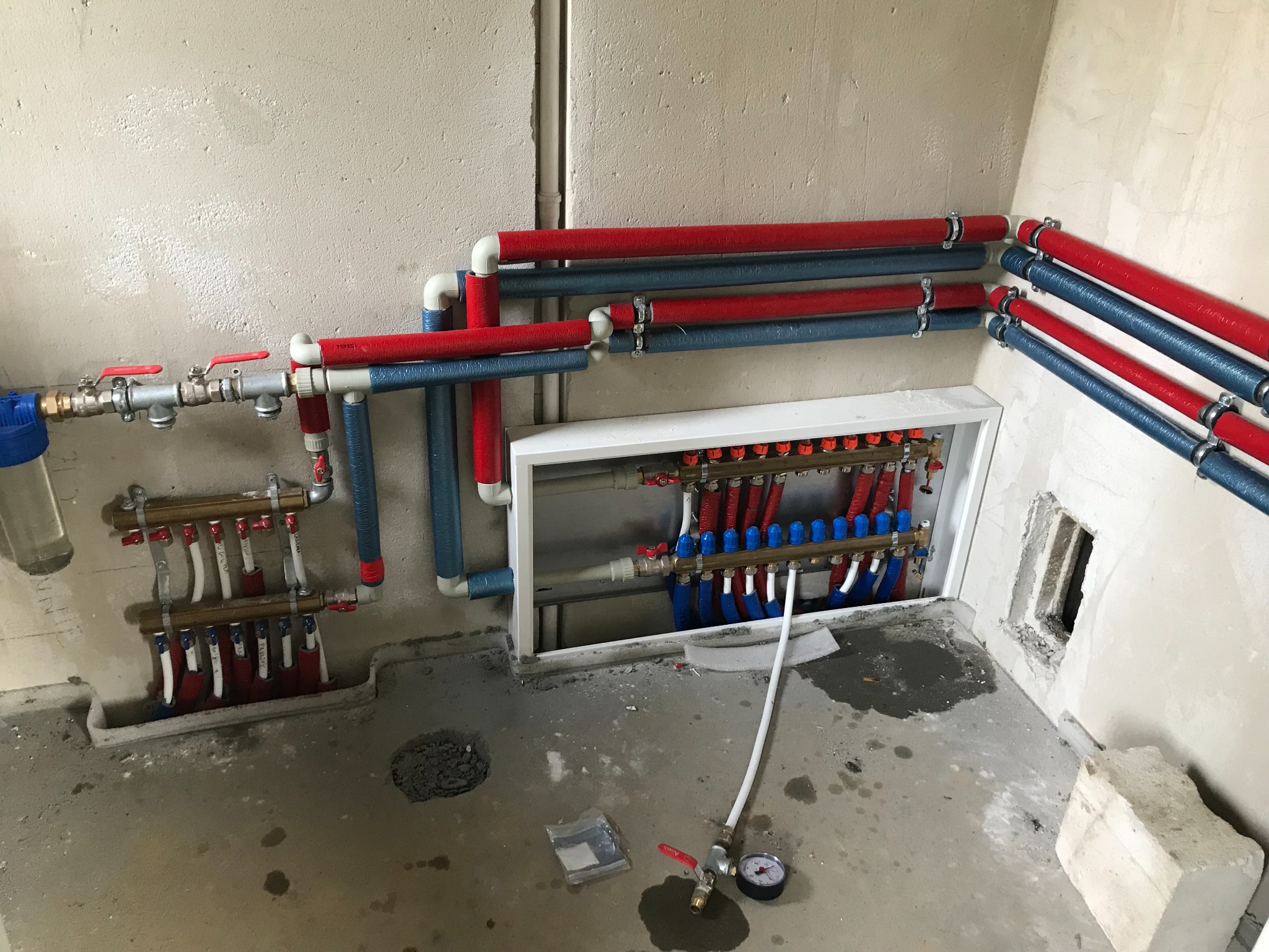 Rozdzielacz centralnego ogrzewania z rurami w izolacji czerwonej i niebieskiej, manometr na podłodze, surowy stan wykończenia ścian i posadzki.