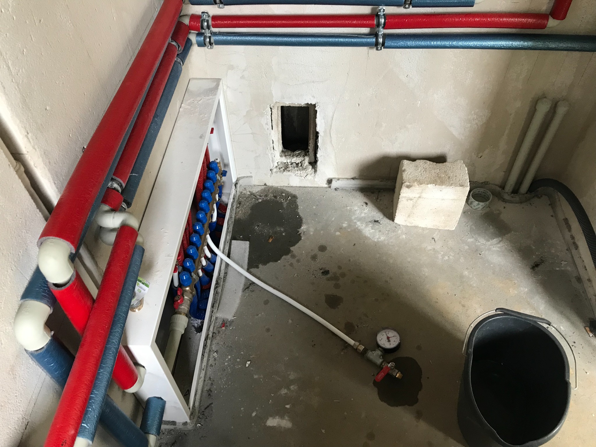 Rozdzielacz instalacji centralnego ogrzewania w szafce, rury grzewcze w izolacji czerwonej i niebieskiej, manometr na podłodze, surowy stan wykończenia pomieszczenia.