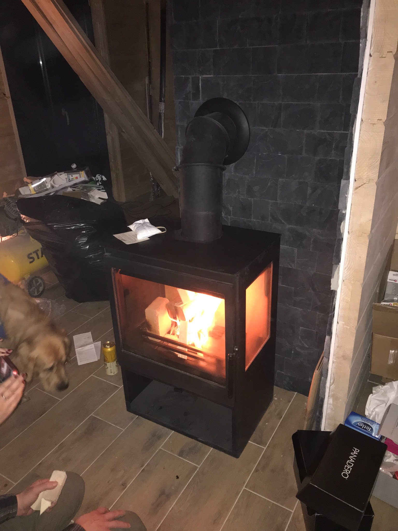 Czarny piec wolnostojący z palącym się w środku drewnem, podłączony do komina z czarnej blachy. W tle ściana wyłożona czarnym kamieniem oraz prowizoryczne wykończenie wnętrza. Na podłodze widoczne...