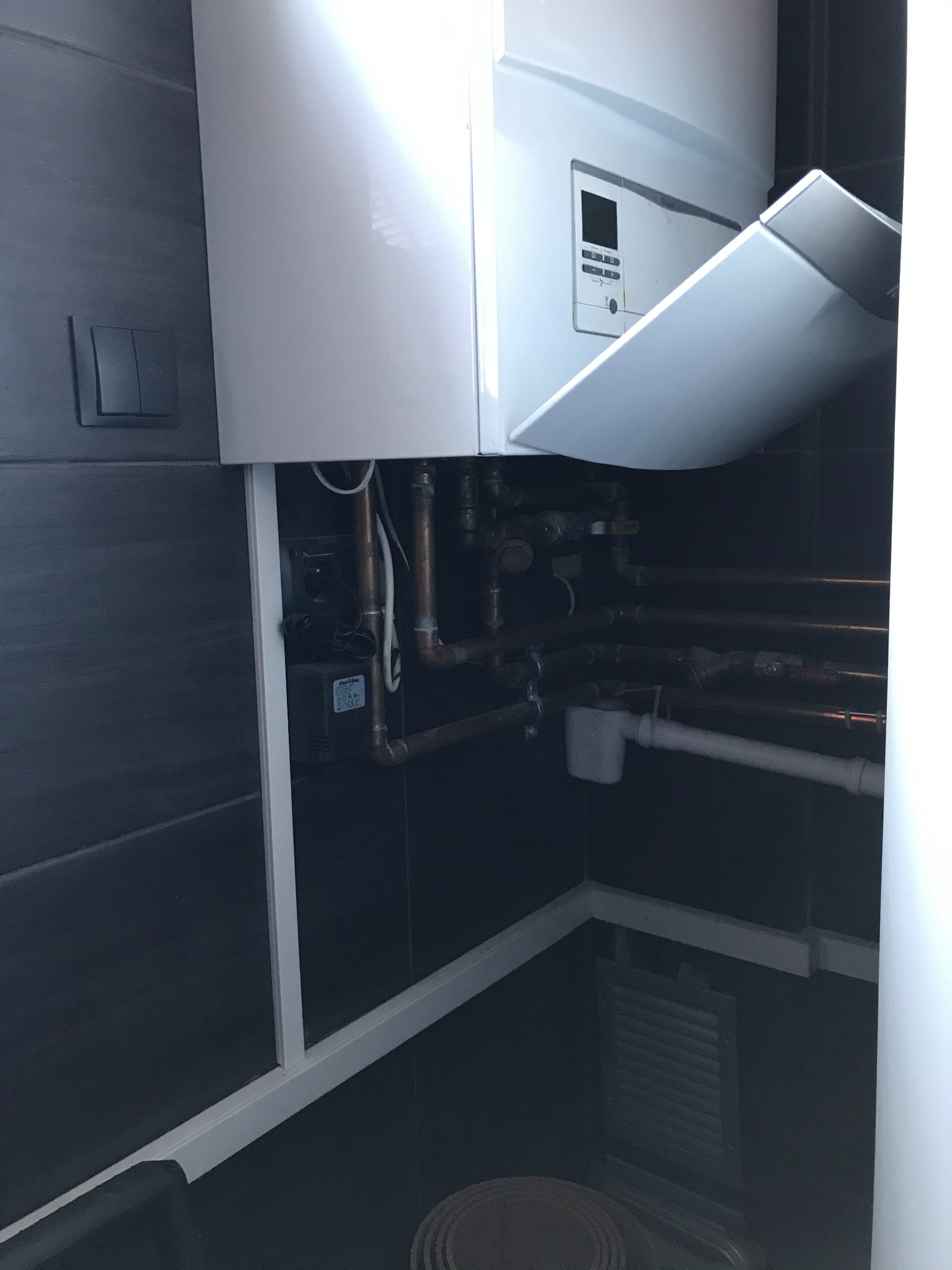 Biały piec gazowy z otwartą klapą, widoczne miedziane rury instalacji w ciemnej wnęce wyłożonej ciemnymi płytkami.
