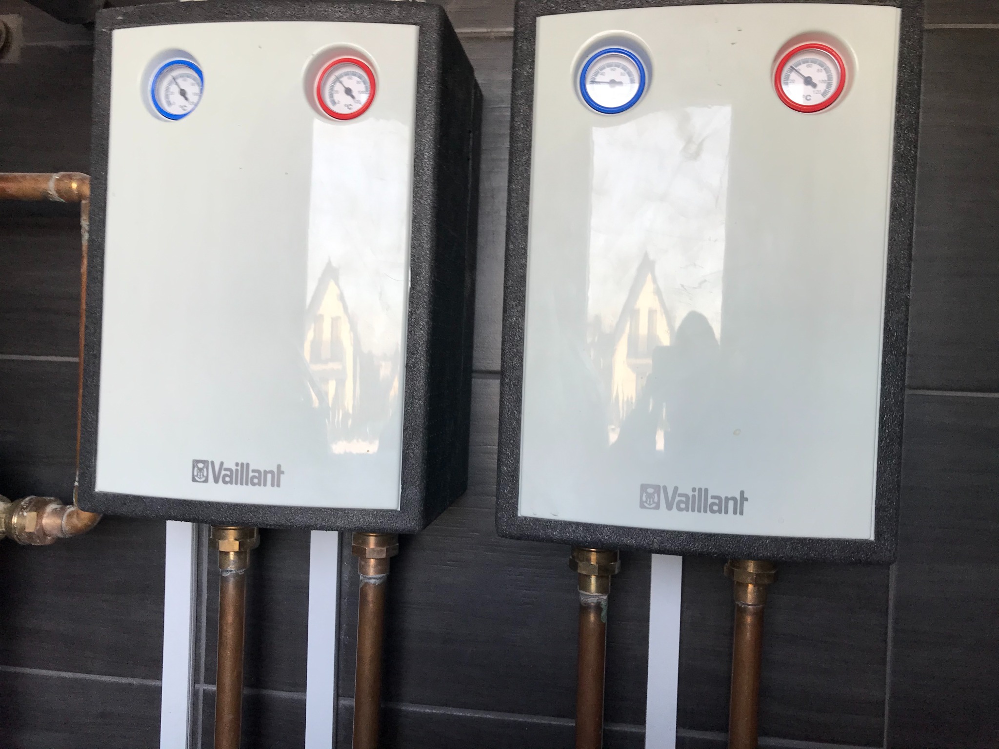 Dwa białe, zaizolowane piece gazowe marki Vaillant z termometrami, podłączone miedzianymi rurami na tle ciemnoszarej ściany z płytek.