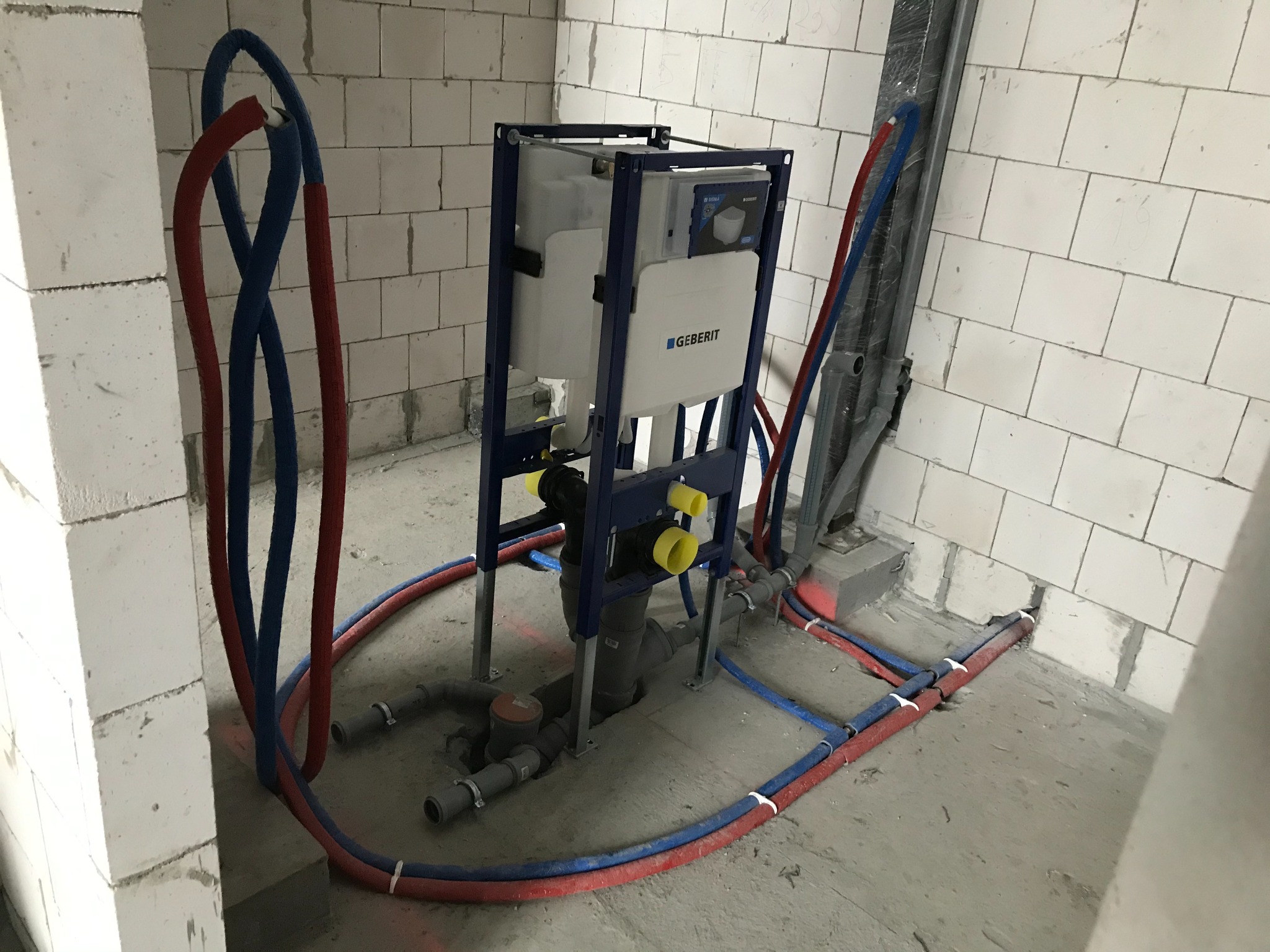 Stelaż podtynkowy Geberit z podłączonymi rurami wodnymi w czerwono-niebieskiej izolacji, widoczne elementy instalacji kanalizacyjnej w surowym stanie, ściany z białych bloczków.